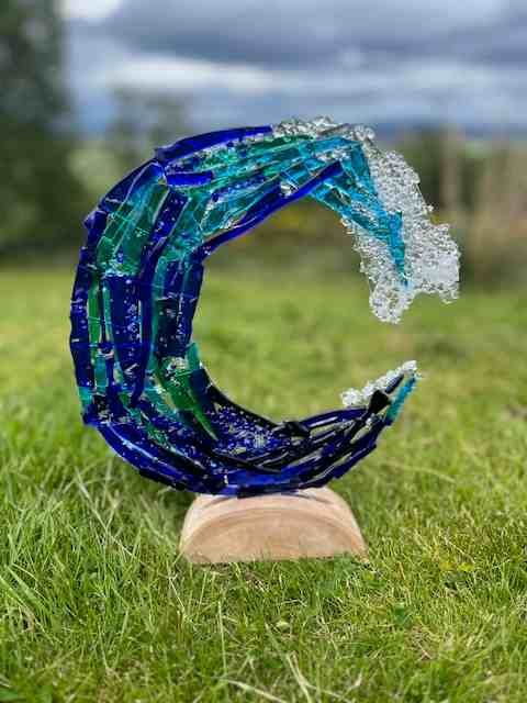 A blue and teal fused glass wave sculpture on a wooden stand, made in the Highlands